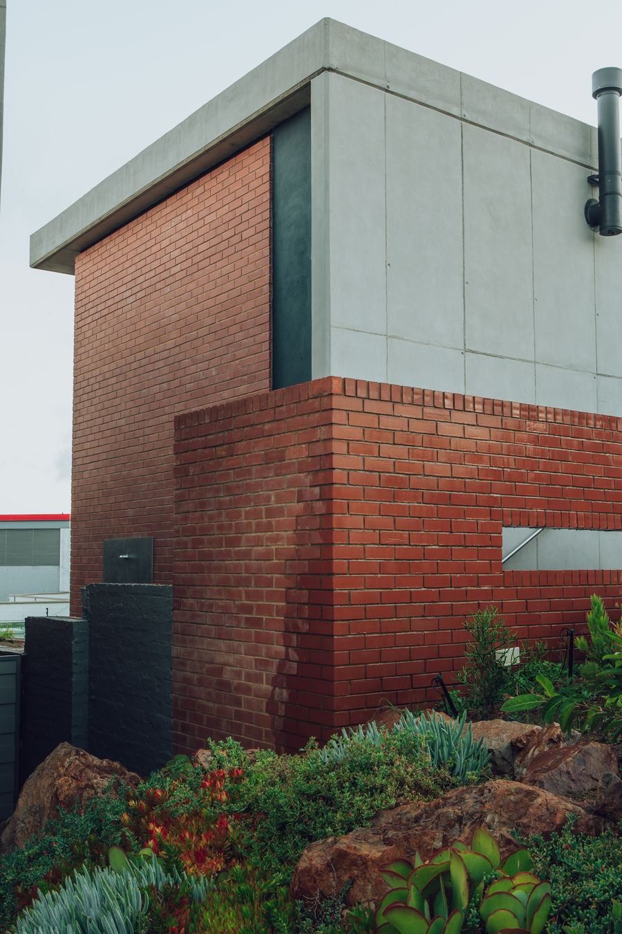 Corner of a house with a red brick base and a concrete upper volume, set among indigenous plants.