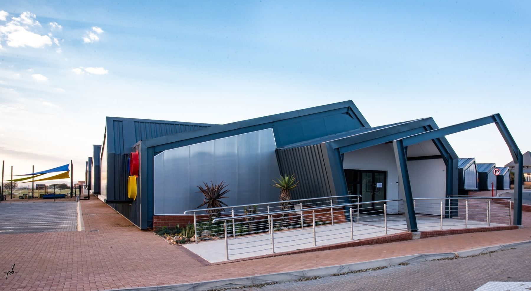 Early childhood development centre with a pitched steel-clad facade and coloured porthole windows, seen from the entrance forecourt.