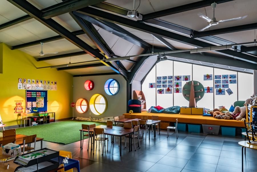 Classroom interior with exposed steel roof trusses, a reading bench and coloured round windows.