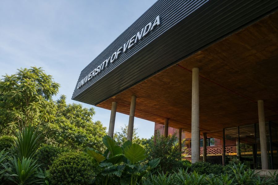 Timber soffit and columns beneath the raised roof, among trees, with the University of Venda name on the cladding.