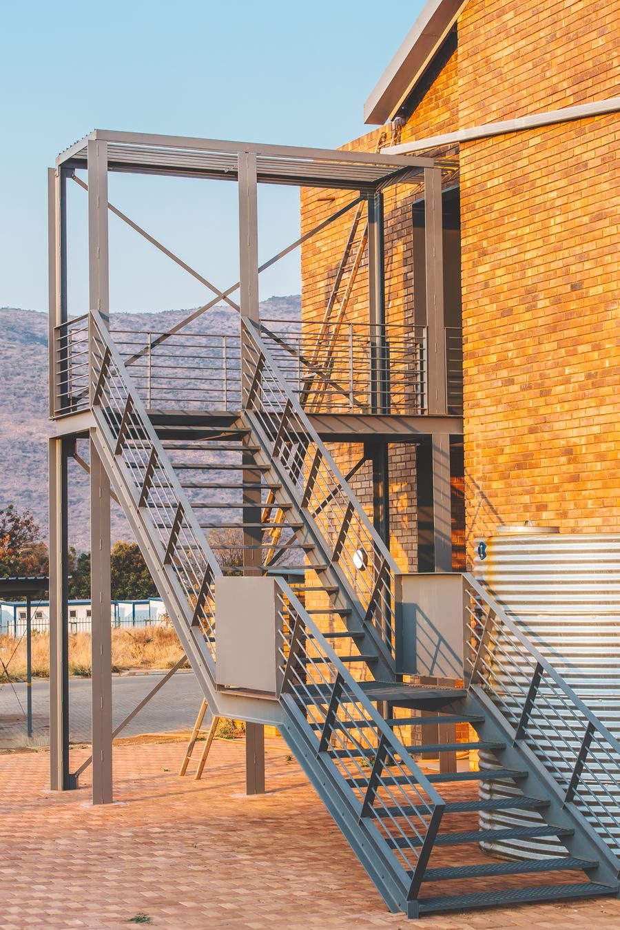 Steel external stair beside a face-brick unit and a water tank, in low sun.